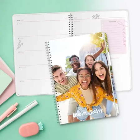 Personalisierter Schülerkalender mit Spiralbindung und individuellem Fotocover 'Mein Jahr', liegend vor einem geöffneten Kalender mit Wochenansicht auf mintgrünem Hintergrund mit Schreibwaren.