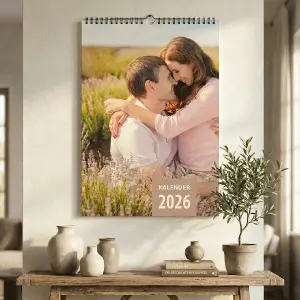Ein personalisierter Wand-Fotokalender 2026 im A3 Hochformat, hängend über einer rustikalen Holzkommode. Das Deckblatt zeigt ein glückliches Paar in einem Lavendelfeld.