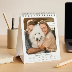 Tischkalender mit Spiralbindung auf einem Holzschreibtisch, platziert neben einem Laptop und Schreibwaren. Das Kalenderblatt zeigt die Woche vom 29. Dezember bis 4. Januar sowie ein Foto eines glücklichen Paares, das einen großen weißen Hund umarmt.