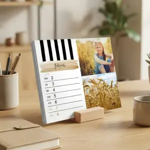 Tischkalender mit Holzsockel auf einem Schreibtisch, der die Februarwoche vom 2. bis 8. zeigt. Die Seite enthält ein Foto eines Mädchens im Gras und ein Bild eines Weizenfeldes, verziert mit schwarz-weißen Streifen und einem goldenen Pinselstrich.